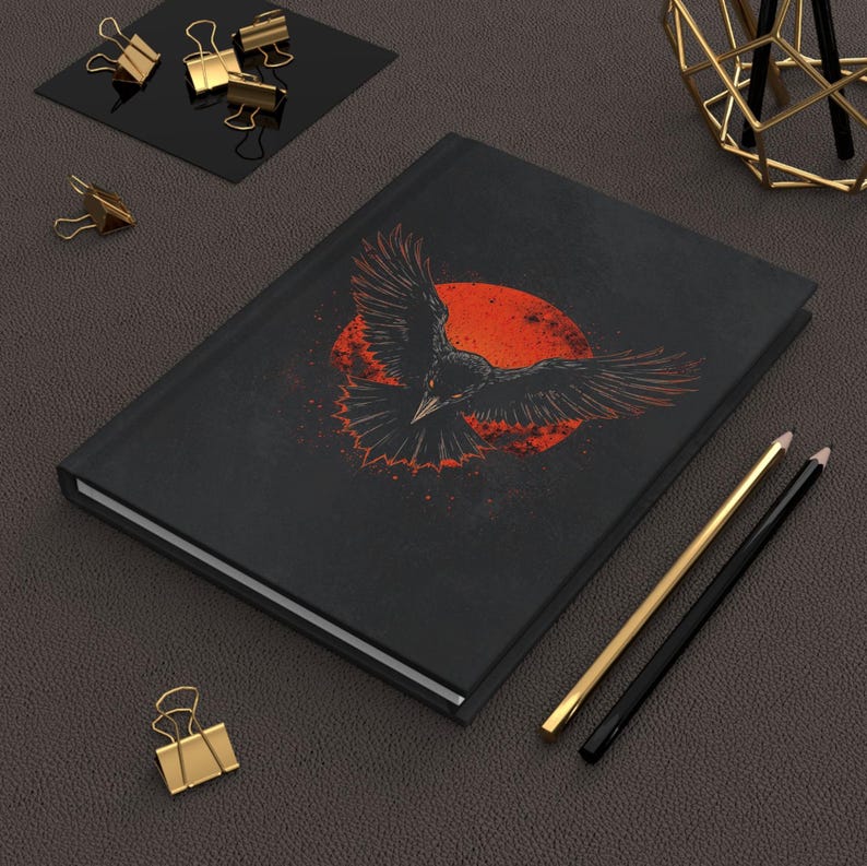 May include: Black journal featuring a raven design with outstretched wings against a red sun. The cover art is detailed with a distressed, vintage aesthetic. Gold and black pencils and binder clips are also visible.