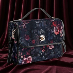 May include: A dark blue handbag with a floral pattern featuring red and pink flowers and green leaves. The bag has a top handle, a detachable shoulder strap, and a silver clasp. White stitching outlines the bag.