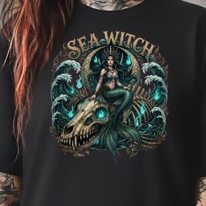 May include: Black t-shirt featuring a detailed illustration of a mermaid with teal hair and a green tail, seated on a skull. The design includes ocean waves and the text "SEA WITCH" in gold lettering.