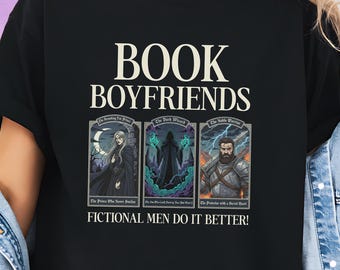May include: Black t-shirt with the text "BOOK BOYFRIENDS" in white. Below the text are three fantasy character illustrations with text: "The Prince Who Never Smiles", "The Dark Wizard", and "The Protector with a Secret Heart". The text "FICTIONAL MEN DO IT BETTER!" is below the illustrations.