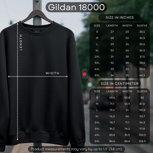 May include: A black Gildan 18000 sweatshirt hangs on a metal rack. The image includes a size chart in inches and centimeters, detailing length, width, and sleeve measurements for sizes S to 5XL. The product measurements may vary by up to 1.5 inches.