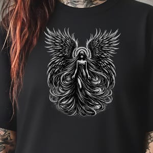 Könnte beinhalten: Schwarzes T-Shirt mit einer schwarz-weißen Illustration eines Engels. Der Engel hat große, detaillierte Flügel und langes, fließendes Haar. Ein Heiligenschein umgibt den Kopf des Engels, und das Design ist zentriert.