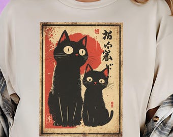 Op de afbeelding: Beige T-shirt met een vintage-stijl grafische afbeelding van twee zwarte katten met grote ogen, tegen een rode en beige achtergrond. Japanse tekst is zichtbaar in het ontwerp. De katten zijn in een gestileerd, illustratief ontwerp.