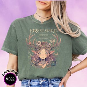 May include: Moss green t-shirt with a graphic of a forest spirit. The design features a woman with antlers, a floral headdress, and butterflies. The words "FOREST SPIRIT" are printed above the graphic. The shirt also has the words "COMFORT COLORS" and "MOSS" printed on it.