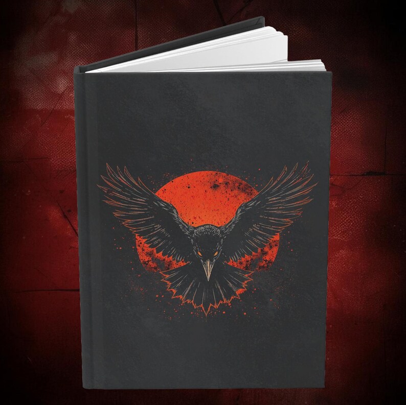 May include: A black journal with a detailed illustration of a raven with outstretched wings against a red sun. The raven design is in shades of black and orange, with white-paged interior. The journal is closed, with the pages slightly visible.