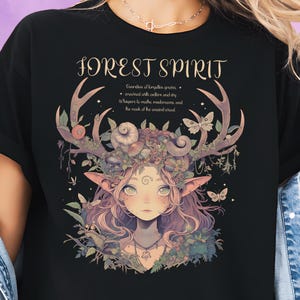 Könnte beinhalten: Schwarzes T-Shirt mit einer Fantasy-Illustration eines Waldgeistes. Das Design umfasst Geweihe, Blumen und Schmetterlinge, mit dem Text "FOREST SPIRIT" darüber. Zusätzlicher Text darunter. Ein skurriles, von der Natur inspiriertes Design.