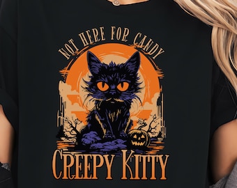 Może przedstawiać: Czarny t-shirt z motywem Halloween. Projekt przedstawia czarnego kota z pomarańczowymi oczami i napisem "NOT HERE FOR CANDY" nad nim. Poniżej znajduje się napis "CREEPY KITTY". Tło obejmuje pomarańczowe słońce i dynię.