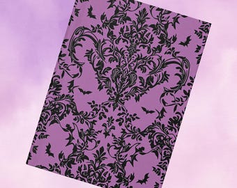 May include: A lavender notebook with a black damask pattern and bat silhouettes. The cover features a detailed floral design with swirling leaves and stems, creating a gothic or Halloween theme. The notebook is rectangular and slightly angled.