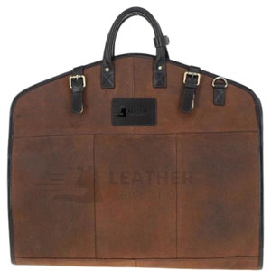 May include: Brown leather garment bag with black straps and buckles. The bag has a handle and a label that says "Leather by Artisans".