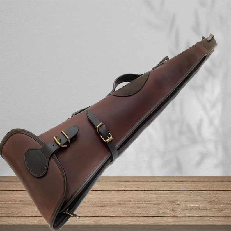 May include: A brown leather gun case with two straps and buckles. The case has a dark brown leather flap with a decorative detail on the front.