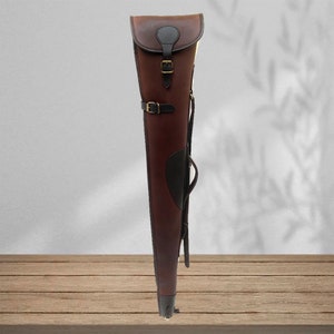 May include: A brown leather gun case with a black leather flap and a brass buckle. The case has a shoulder strap and a carrying handle.