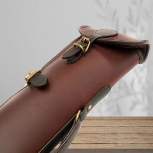 May include: A brown leather gun case with a black strap and brass buckles. The case has a flap with a buckle closure.