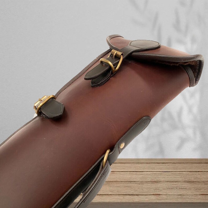 May include: A brown leather gun case with a black strap and brass buckles. The case has a flap with a buckle closure.