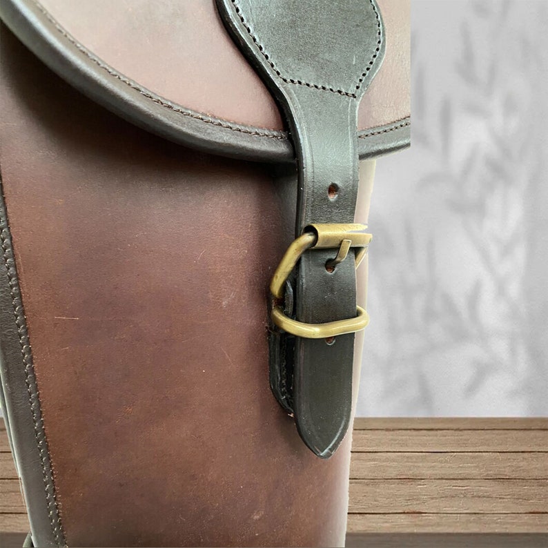 May include: A brown leather bag with a black strap and a brass buckle. The bag has a flap with a stitched edge.