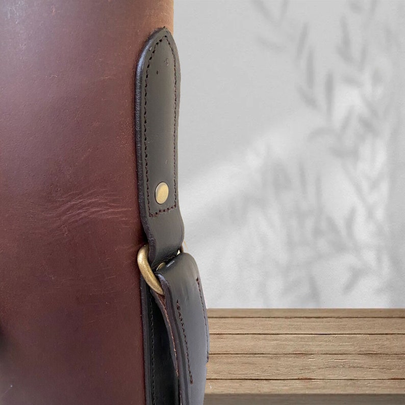 May include: Close-up of a brown leather bag with a black leather strap and a brass buckle. The bag has a textured surface and a simple design.