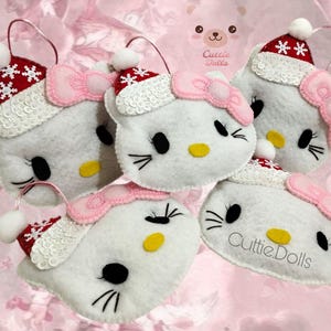 May include: Five white plush ornaments shaped like a cartoon cat face, each with a pink bow, black whiskers, and a yellow nose. They wear red and white Santa hats with snowflake details. The ornaments have a loop for hanging and the text "Cuttie.Dolls".