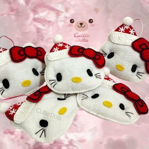 May include: Five white felt ornaments shaped like a cartoon cat with a red bow, black eyes, and a yellow nose. Each ornament has a white and red Santa hat with a white pom-pom. The ornaments are from Cuttie Dolls.