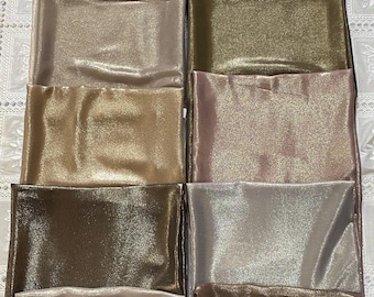May include: A collection of folded, shimmering fabric squares in various colors, including gold, bronze, and black. The fabric has a glossy, reflective surface, suggesting a satin or silk-like material. The arrangement showcases the range of available shades.