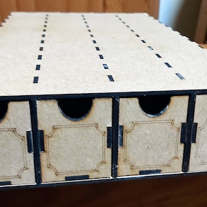 Può includere: Una scatola di legno con quattro cassetti estraibili. La scatola è realizzata in legno marrone chiaro con dettagli neri. La parte superiore ha linee distanziate uniformemente. Ogni cassetto ha un'area etichetta decorativa.