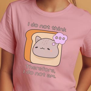 Puede incluir: Camiseta rosa con un gato de dibujos animados sobre una tostada y el texto "I do not think, Therefore, I do not am." La camiseta tiene mangas cortas y cuello redondo. El color es rosa.
