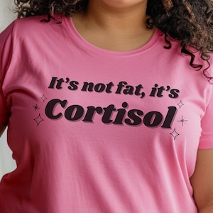 Könnte beinhalten: Ein pinkfarbenes T-Shirt mit dem Aufdruck "It's not fat, it's Cortisol" auf der Vorderseite. Der Text ist schwarz und in einer stilisierten Schriftart gehalten.