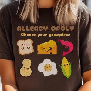 Può includere: Maglietta marrone con il testo "Allergy-Opoly Scegli il tuo pezzo di gioco" e immagini di cartoni animati di una fetta di pane, un pezzo di formaggio, un gambero, un arachide, un uovo e una pannocchia di mais.