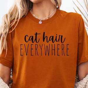 May include: A woman wearing a burnt orange t-shirt with the text "cat hair everywhere" printed on it.