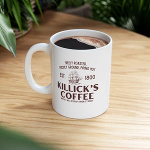 May include: White ceramic coffee mug filled with dark coffee. The mug features the text "FREELY ROASTED, FRESHLY GROUND, PIPING HOT, EST. 1800, KILLICK'S COFFEE, WHICH IT WILL BE READY WHEN IT'S READY" in brown. The mug has a curved handle.