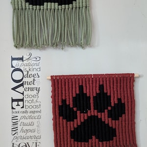 May include: Two macrame wall hangings with a black and white geometric design. The top one is green and the bottom one is red. The red one has the text "LOVE ALWAYS" and "DOVE NEVER FAILS" printed on the wall behind it.