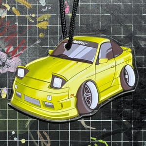 May include: Yellow car-shaped air freshener with black string. The car has a cartoon design with white wheels and a black tire. The text "ApeNK" is on the windshield. The car is a coupe with a spoiler.