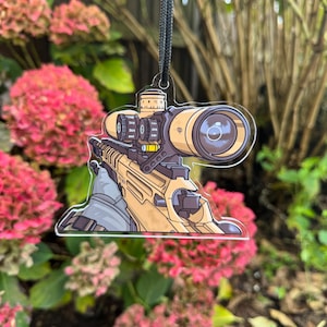 Puede incluir: Un rifle de color beige con una mira telescópica y un cordón negro adjunto. El rifle está dibujado en un estilo de dibujos animados y está en un llavero de acrílico transparente.