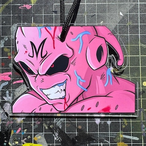 May include: A pink acrylic charm depicting a cartoon character with a sinister grin, sharp teeth, and red eyes. The character has blue vein-like markings and the letter "M" on its forehead. A black cord is attached for hanging.