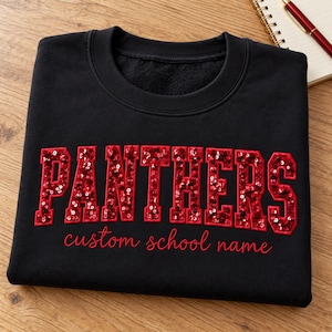 May include: A black crewneck sweatshirt featuring the word PANTHERS in large, red, sequined block letters. Below the word, the phrase custom school name is embroidered in red cursive script. The garment has a ribbed collar and long sleeves.