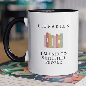 Puede incluir: Taza de cerámica blanca con asa negra y borde negro. La taza tiene el texto "LIBRARIAN" y "I'M PAID TO SHHHHHHH PEOPLE" impreso en ella. Hay una pila de libros coloridos ilustrados en la taza.