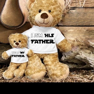 May include: Two brown teddy bears wearing white t-shirts. The larger bear's shirt says "I AM HIS FATHER" and the smaller bear's shirt says "HE IS MY FATHER".