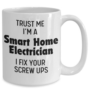 Op de afbeelding: Witte keramische mok met een gebogen handvat. De mok heeft zwarte tekst met de tekst "TRUST ME I'M A Smart Home Electrician I FIX YOUR SCREW UPS".
