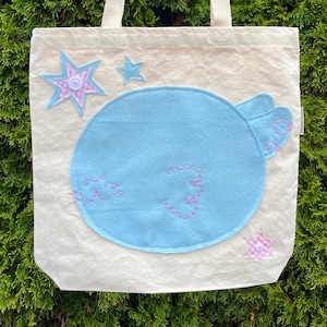 May include: A cream-colored tote bag featuring a large, light blue circular felt applique of a sleeping animal face with pink embroidered details. The design includes two light blue felt stars and one pink and white checkered star with a white button center.