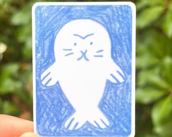 May include: A rectangular sticker featuring a white seal illustration with a blue border. The seal has a simple, cartoonish design with a frowning expression. The sticker is held by a hand against a backdrop of green foliage.