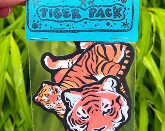 May include: A package of tiger stickers. The stickers feature orange and black tiger designs with white accents. The package is held by a hand and has a blue header with the words "TIGER PACK" printed on it.
