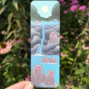 May include: A rectangular bookmark featuring a vertical illustration of Japanese macaques in a hot spring. The design includes a light blue sky with a pale sun, dark green trees, grey rocks, a blue waterfall, and several monkeys with pink faces.