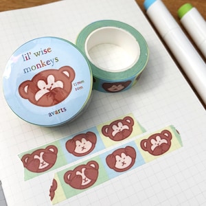Puede incluir: Dos rollos de cinta adhesiva decorativa con diseño de mono, etiquetados como "lil' wise monkeys" y "avarts". La cinta es azul claro con caras de mono marrón. Dos bolígrafos blancos con puntas azules y verdes también están en la imagen. La cinta mide 1,5 cm de ancho y 10 m de largo.