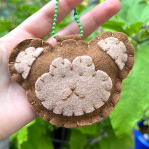 May include: A handmade brown felt bear-shaped ornament with tan accents and embroidered details. The bear has a light brown face and ears, with a stitched nose and mouth. A green ball chain is attached at the top for hanging.