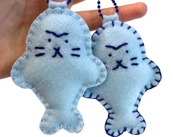 May include: Two light blue felt seal ornaments with dark blue stitched details and facial features. Each seal has a blue chain attached at the top. The ornaments are handmade and have a whimsical, cartoonish design.