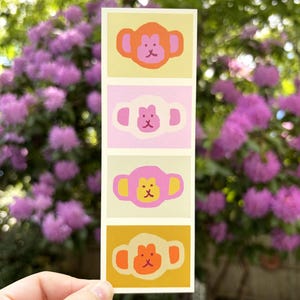 May include: A rectangular sticker featuring four cartoon monkey faces in various colors. Each monkey has a round head, large ears, and a small face. The background colors are yellow, pink, and orange.