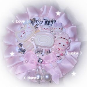 Könnte beinhalten: Pastellrosa dekorative Brosche mit einer geschichteten Schleifenbasis. Die Brosche zeigt verschiedene Charms im Kawaii-Stil, darunter eine Katze, ein Bär und ein Kuchen, verziert mit Strasssteinen und Perlen. Die Texte "Love", "Lucky" und "Happy" sind sichtbar.