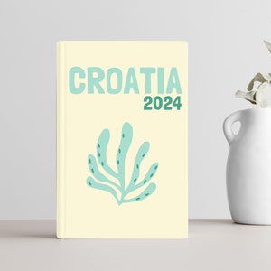Puede incluir: Un libro amarillo con el texto "CROATIA 2024" en letras verdes y azules. Un diseño estilizado de algas marinas azules y verdes está en la portada.