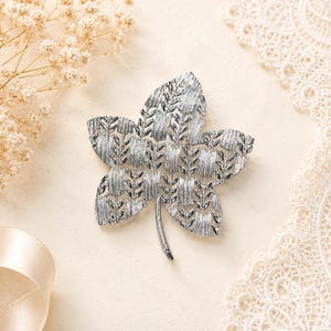 May include: A silver-toned metal brooch shaped like a five-pointed leaf. The surface features a textured, woven-like pattern with alternating vertical and horizontal etched lines, creating a detailed, dimensional appearance with a single stem at the base.
