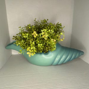 May include: A matte light blue ceramic planter shaped like a conch shell, filled with a dense cluster of small, vibrant green artificial leaves. The planter features a smooth, curved exterior with subtle ridges along the tapered end.