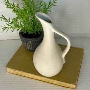 May include: A small, off-white ceramic pitcher with a curved handle and spout. The pitcher sits on a vintage book with a yellowed cover, next to a small potted plant with green foliage. The background is a white wooden surface.