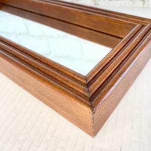 May include: A rectangular wooden framed mirror with a warm brown finish. The frame has a layered design with a smooth, glossy surface. The mirror reflects a white surface.